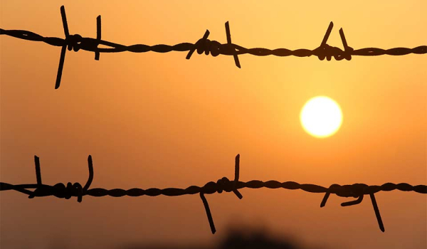 A bleary orange sun is setting, as seen through a barbed wire fence.