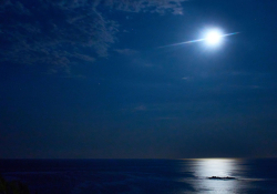 A photograph of a look out at sea, where the moon, just wrapped in gauzy clouds, hovers over a motionless sea below