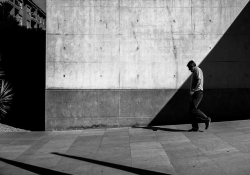 A black and white photo of a man walking down a sloping street in front of a wall transected by shadow