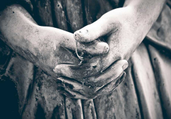 A somber black and white photo of the clasped hands of a statue, darkened by weathering