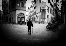 A black and white photo of a man using a cane staring at the buildings in an otherwise empty city