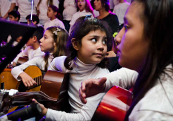 A young girl holding a guitar and surrounded by other children with instruments looks behind her
