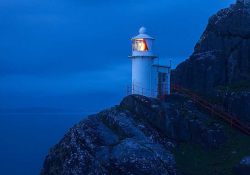 A photograph of a lighthouse perched on the face of a cliff, shrouded in blue dusk