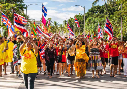 A group of people, clad in brightly colored clothes, march toward the camera waving the Hawai'ian flag