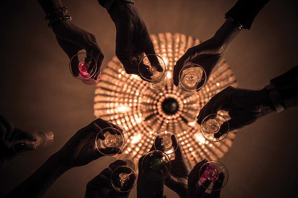 Looking up at a group of hands holding champagne glasses with a chandelier lighted but unfocused in the background.