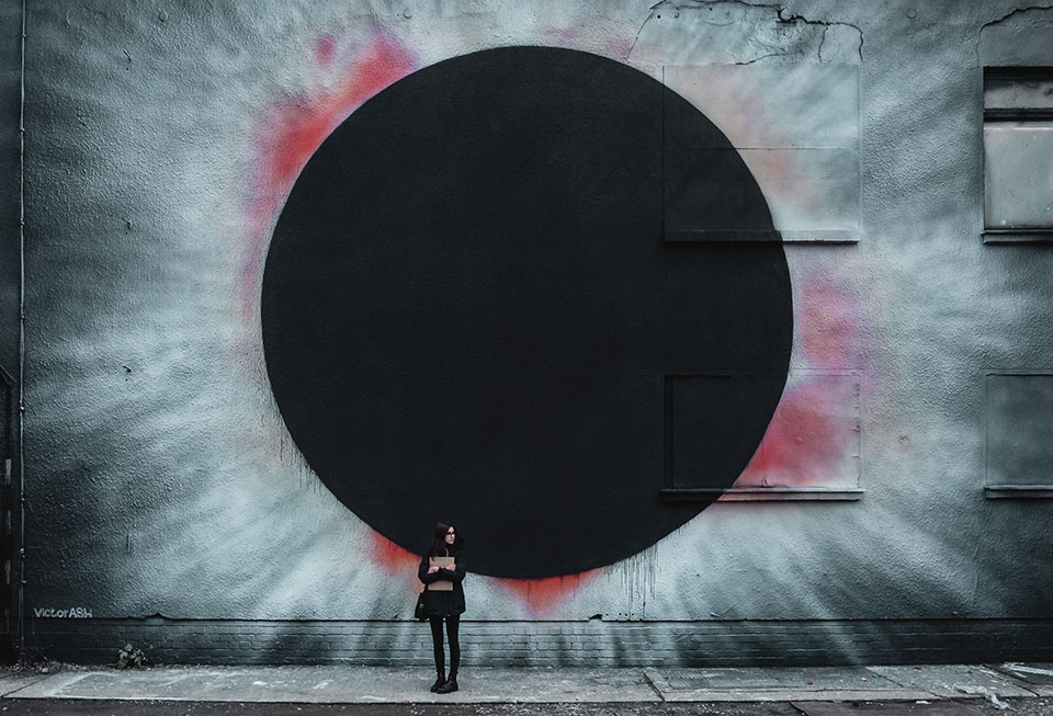 A photograph of a woman standing in front of the wall of an industrial looking building with a spray-painting of a black hole on the side of it