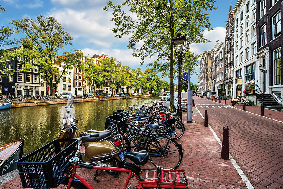 A photograph of a row of bikes parked by a canal that runs parallel to a picturesque city street in Amsterdam