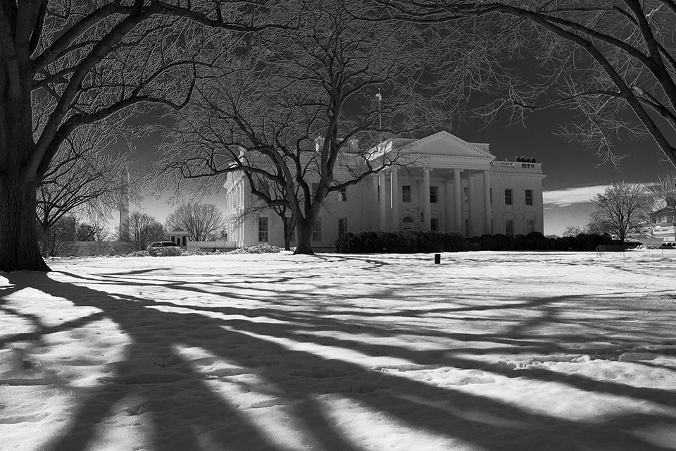 A black and white photo of the White House, seen with long tree shadows lain ominously across the snowy ground
