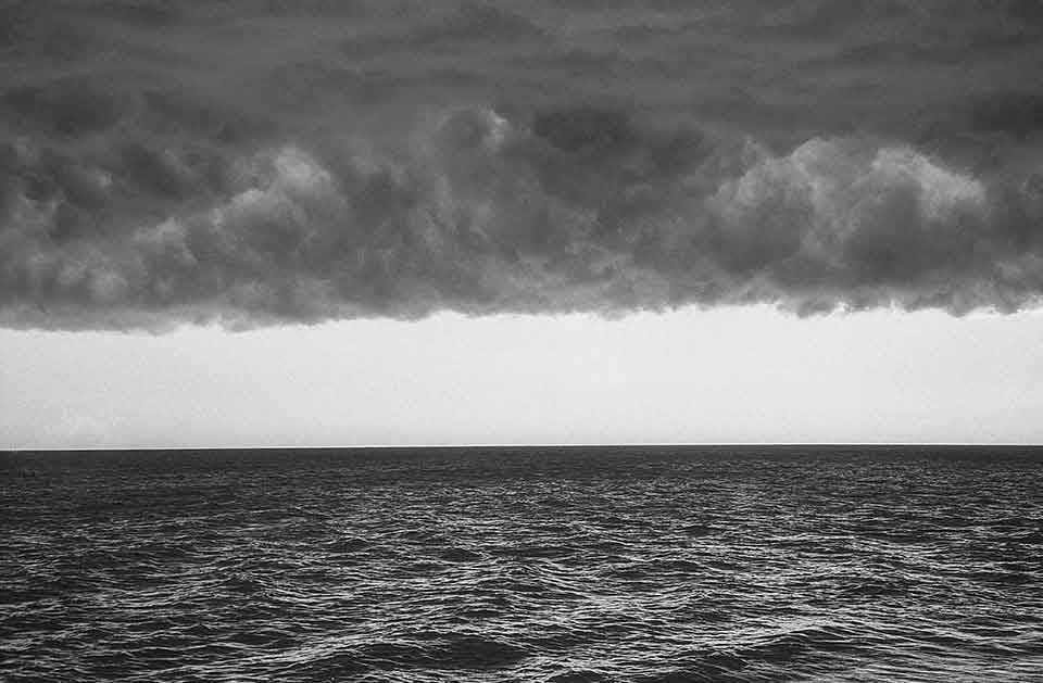 A black and white photo of the sea, dark clouds looming overhead, with the horizon forming the only light between them