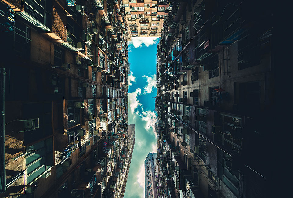 A photo looking upward between two very tall buildings with a blue sky, dotted with clouds, emerging in the thin line between them
