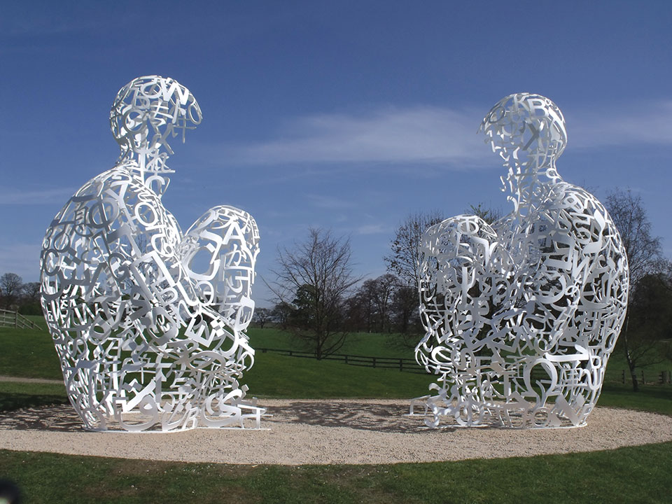 An outdoor sculpture installation featuring two figures seated and facing one another. The figures are composed of calligraphy and are hollow and unfinished.