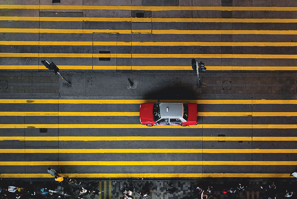 A shot from directly above of a red taxi cab waiting at a street light on a street marked by numerous parallel yellow lines