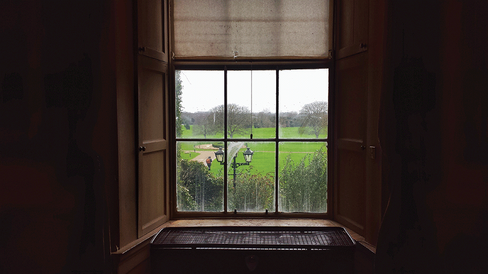 A photograph shot from inside a dark house, looking out a nearby window where greenery rolls out like a lush carpet beneath clouded skies