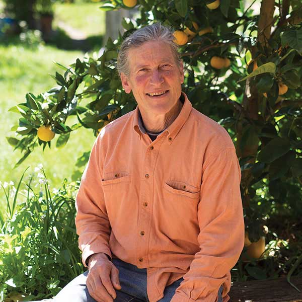 David Holmgren, sitting in the shade of a fruit tree, smiling at the photographer