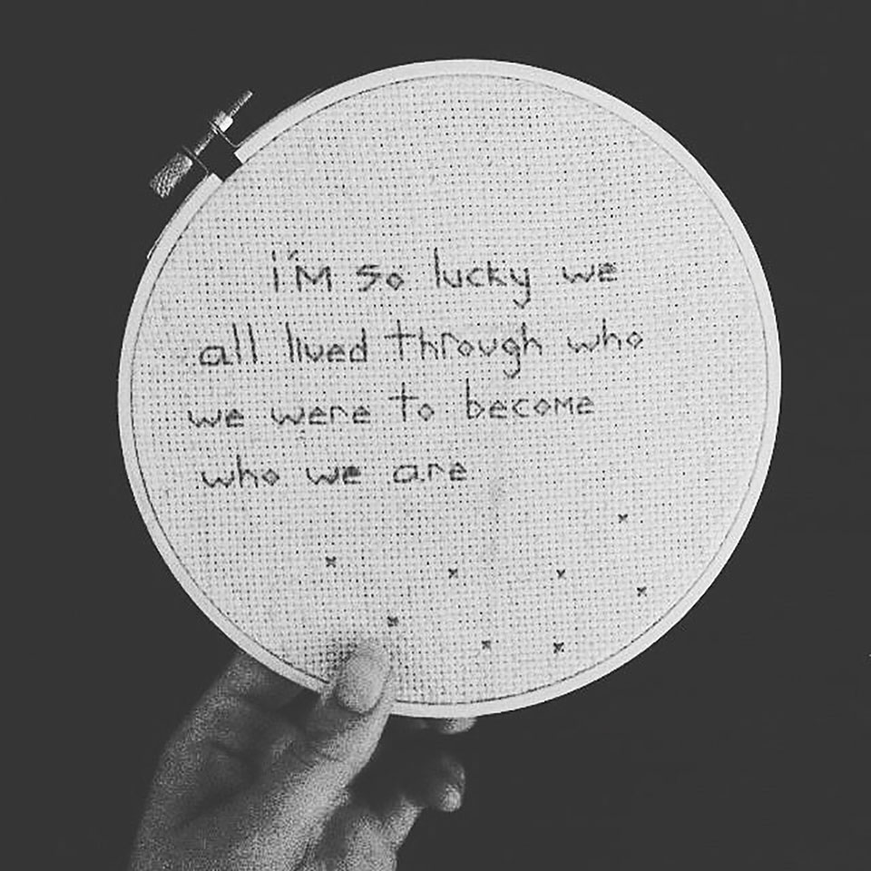 Words needlepointed into cloth on a hoop. The words read I'm so lucky we all lived through who we were to become who we are