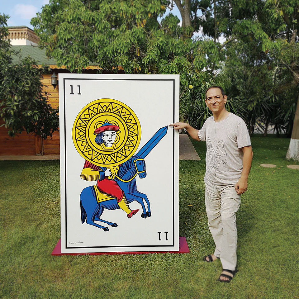 Youssouf Amine Elalamy standing next to a human sized playing card. He points to the tip of a sword.