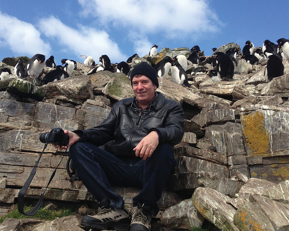 Robin Hemley sits on a outcropping of rock with peguins mingling above him