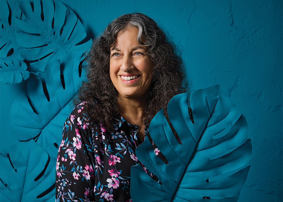 Margarita Engle smiles, holding a broad leaf, painted blue, in front of a blue background