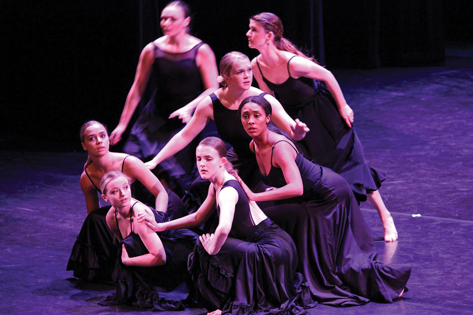 Seven dancers hold a pose on a stage lit in purple