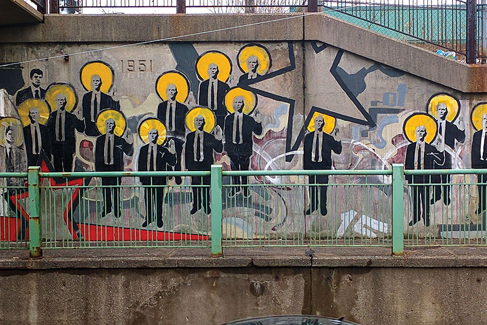 Grafiitti on the side of an off-ramp, dominated by a repeating figure of a man in a suit, sporting a halo like Byzantine iconography