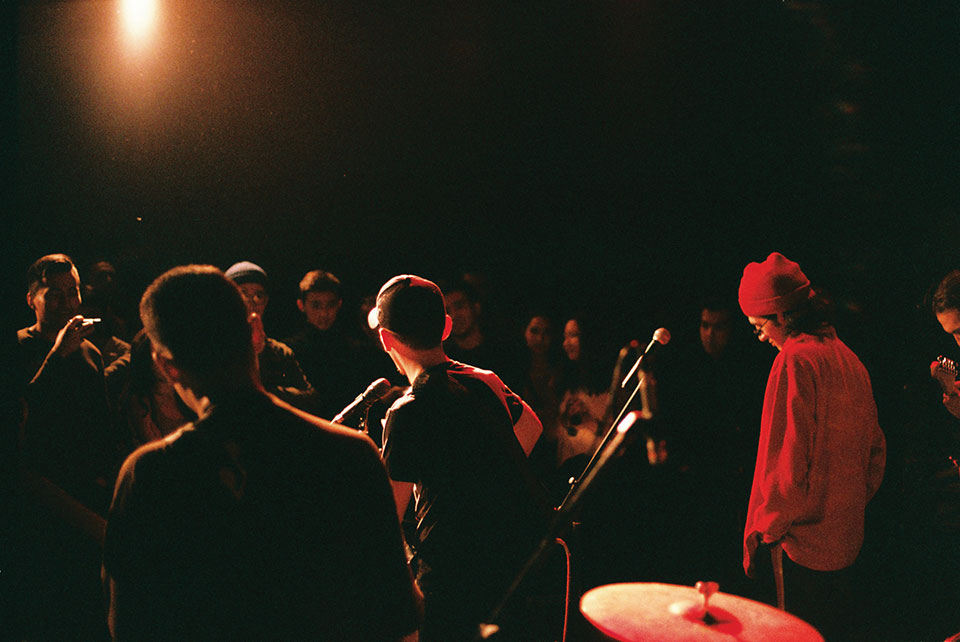 Three musicians stand on a lit stage in a darkened room with the audience standing just inches away