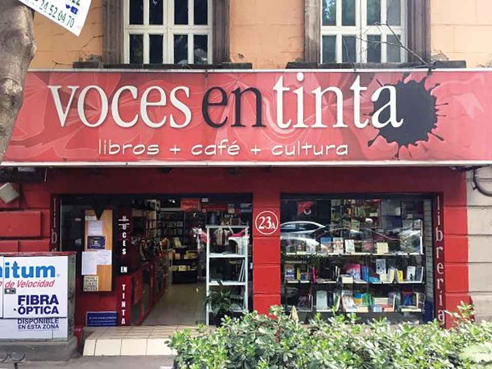 A photograph of a red bookstore front from the street. The sign above reads: Voces en Tinta with a subheading that reads: Libros + Cafe + Cultura