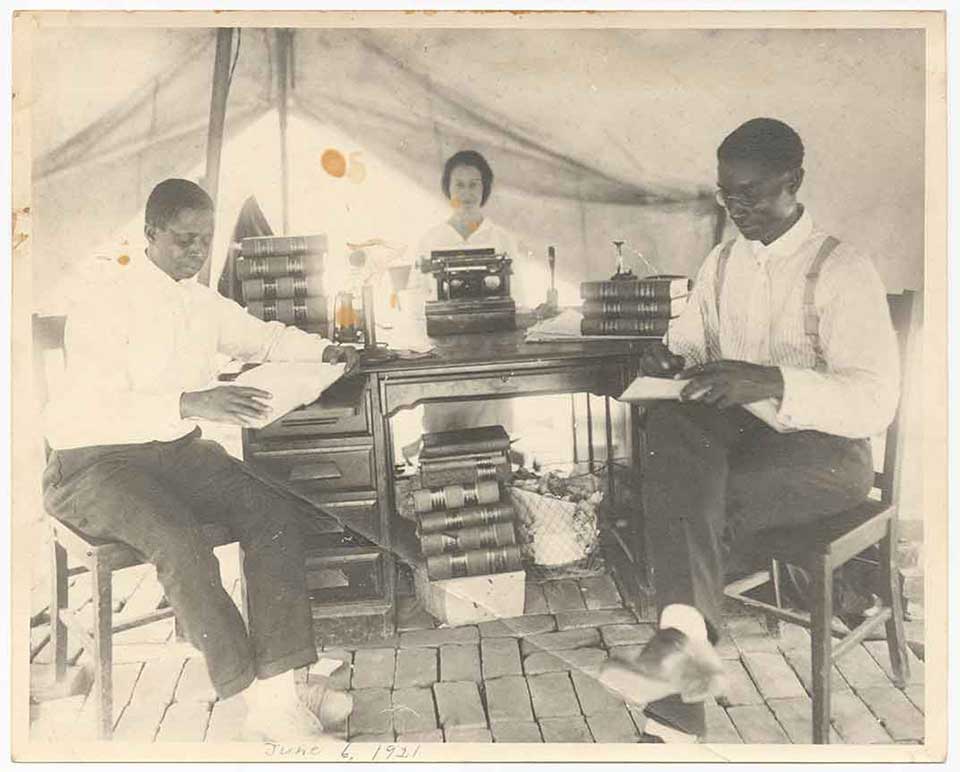 Two African American men seated at a table in a tent. Law books are stacked on the table. A woman stands at the entrance to the tent in the background.