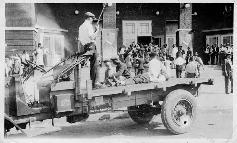 A black and white photograph. In the foreground is an open bed truck. In the bed, a man stands with a shotgun. An African-American man tends to the body of another, laid out on the bed.