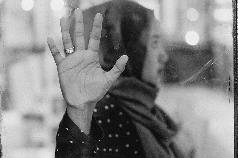 A black and white photograph of a woman, whose head is covered by a scarf, pressing her hand to a damp window between herself and the viewer. Her gaze is away from the viewer.