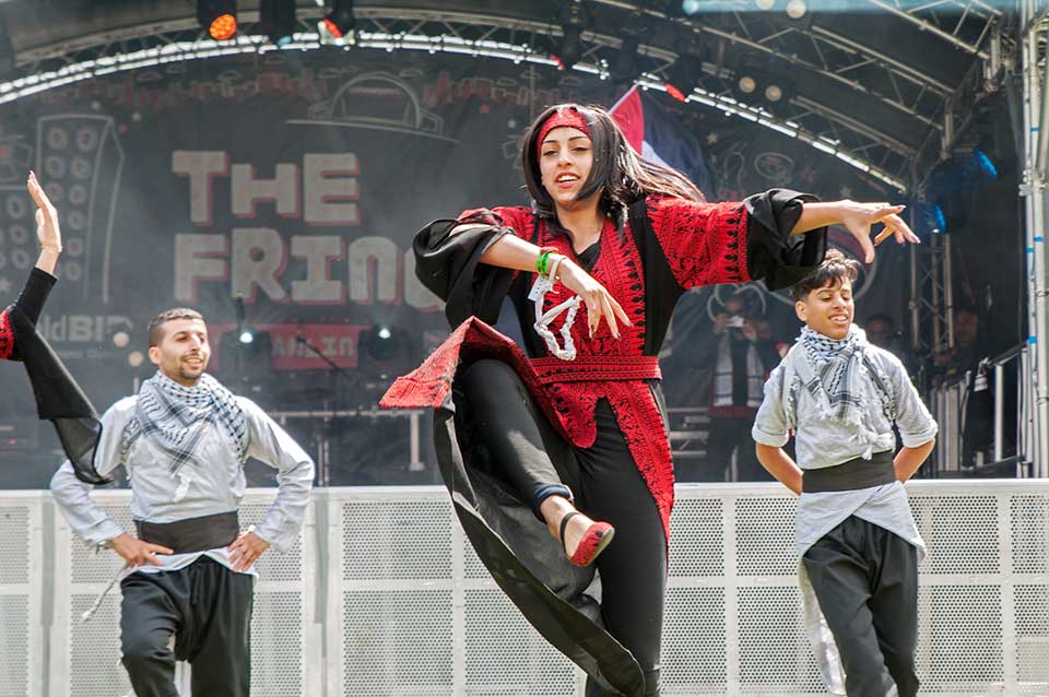 Three dancers on a large outdoor stage. Two male dancers, dressed in grey and black, flank a female dancer, dressed in black and red