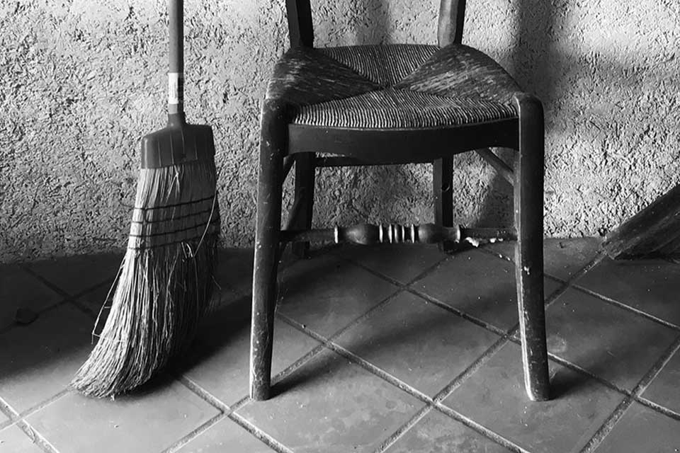 A black and white photograph of the bottom half of a broom and a chair against a rough-hewn wall