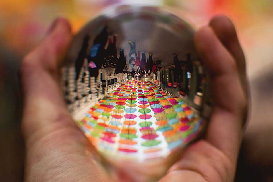 A polished steel ball held in a hand reflects a long table with colored plates on it
