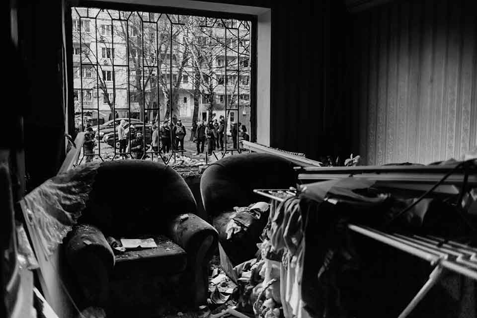 A black and white photograph of two chairs inside a home. Rubble has fallen on them, presumably from a bomb blast