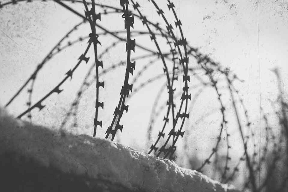 A black and white photograph of rolls of barbed wire affixed above a stone wall