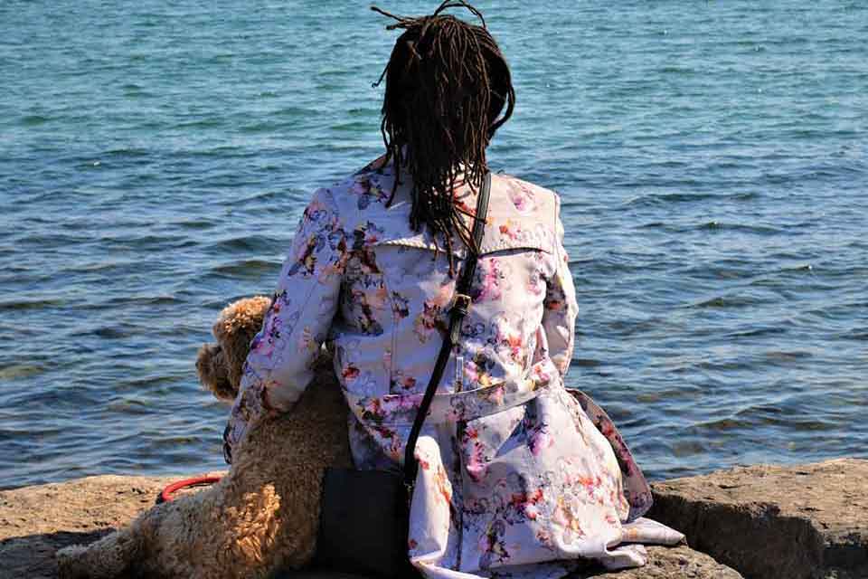 A photograph of a dreadlocked figure sitting with a dog looking out over a body of water