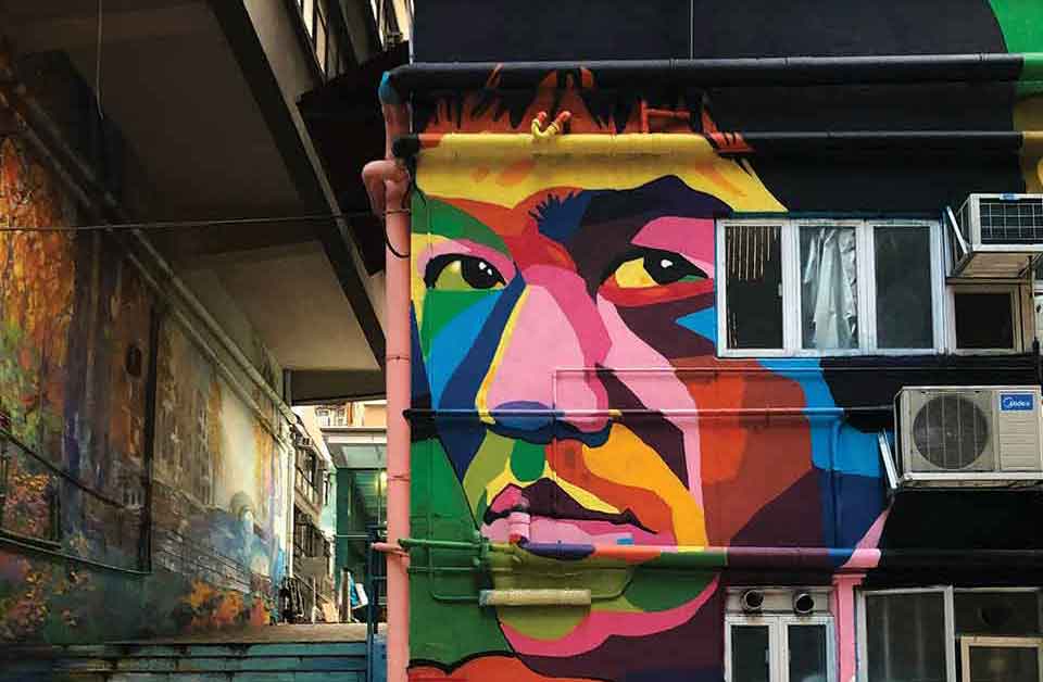 A photograph of a mural on a wall in a crowded urban environment. The mural is of Bruce Lee's face, rendered in bright colors.