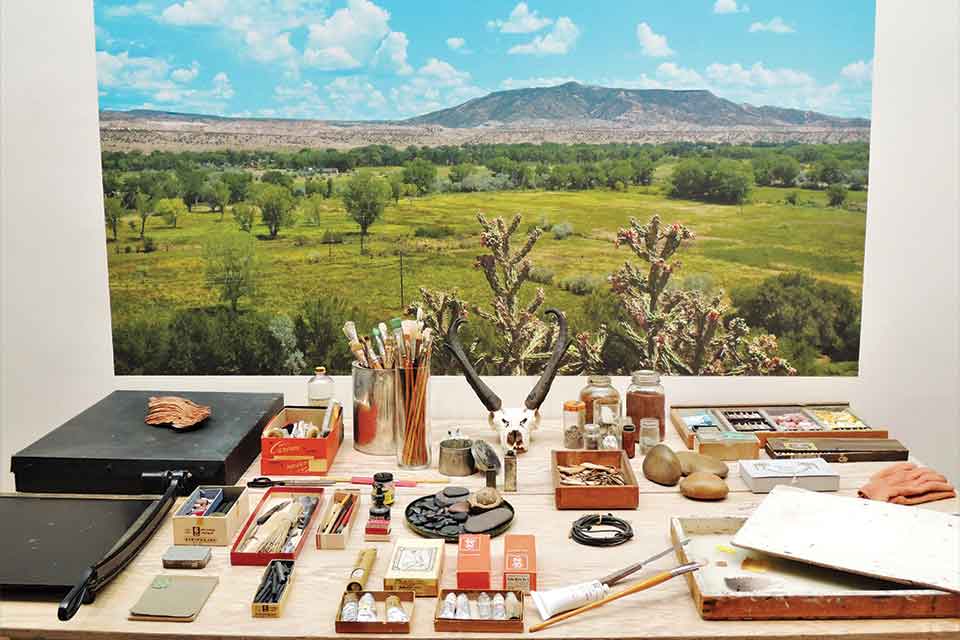 A table full of painting tools with a painting of a New Mexican landscape mounted on the wall behind it