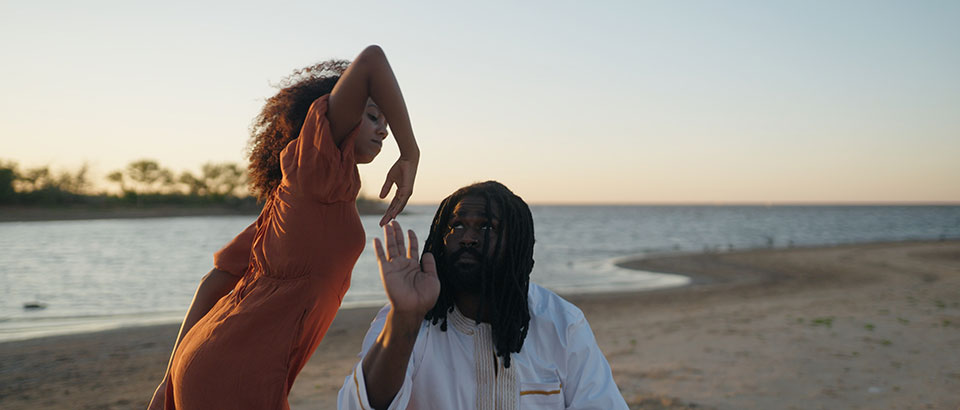 A woman dances expressively near a man who is seated on a beach
