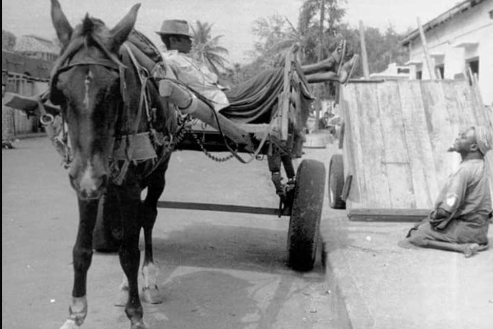 A black and white film still. A man reclines in the back of a wagon pulled by a donkey while another man, sitting on the sidewalk adjacent, looks at him