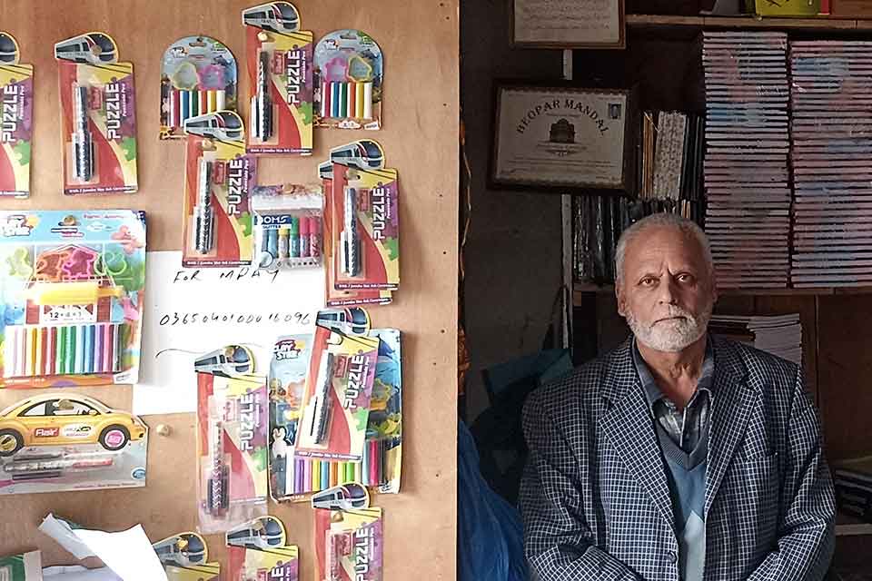 A photograph of an older man standing next to a wall with writing supplies on it