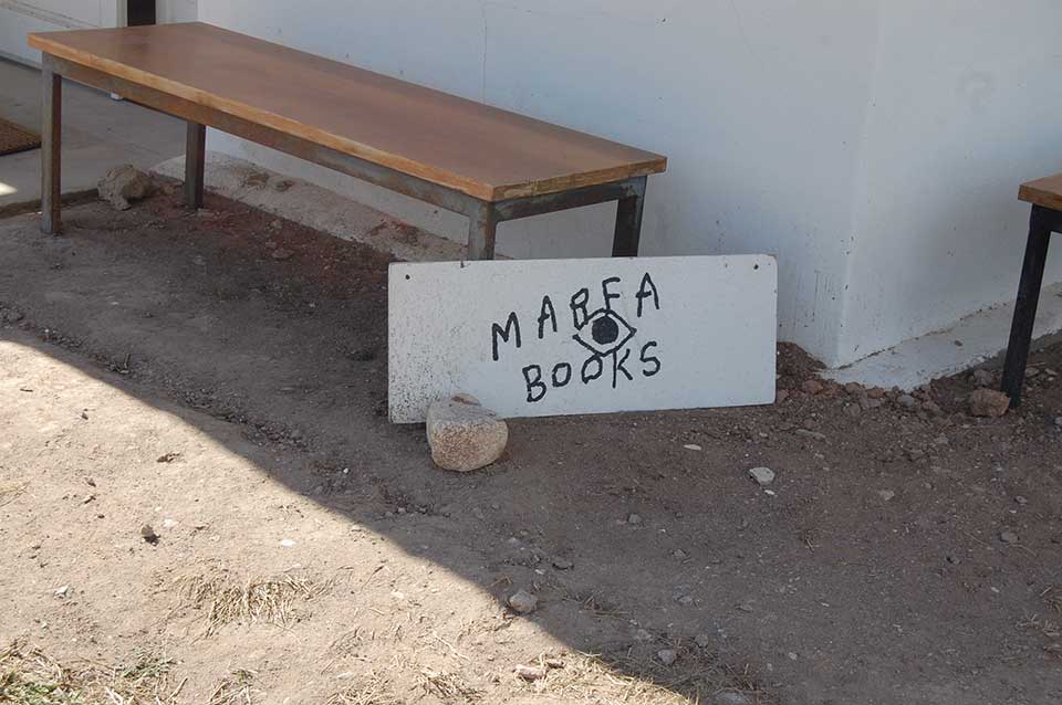 A photograph of a hand-lettered sign propped up against a low wooden bench. Text reads: Marfa Books