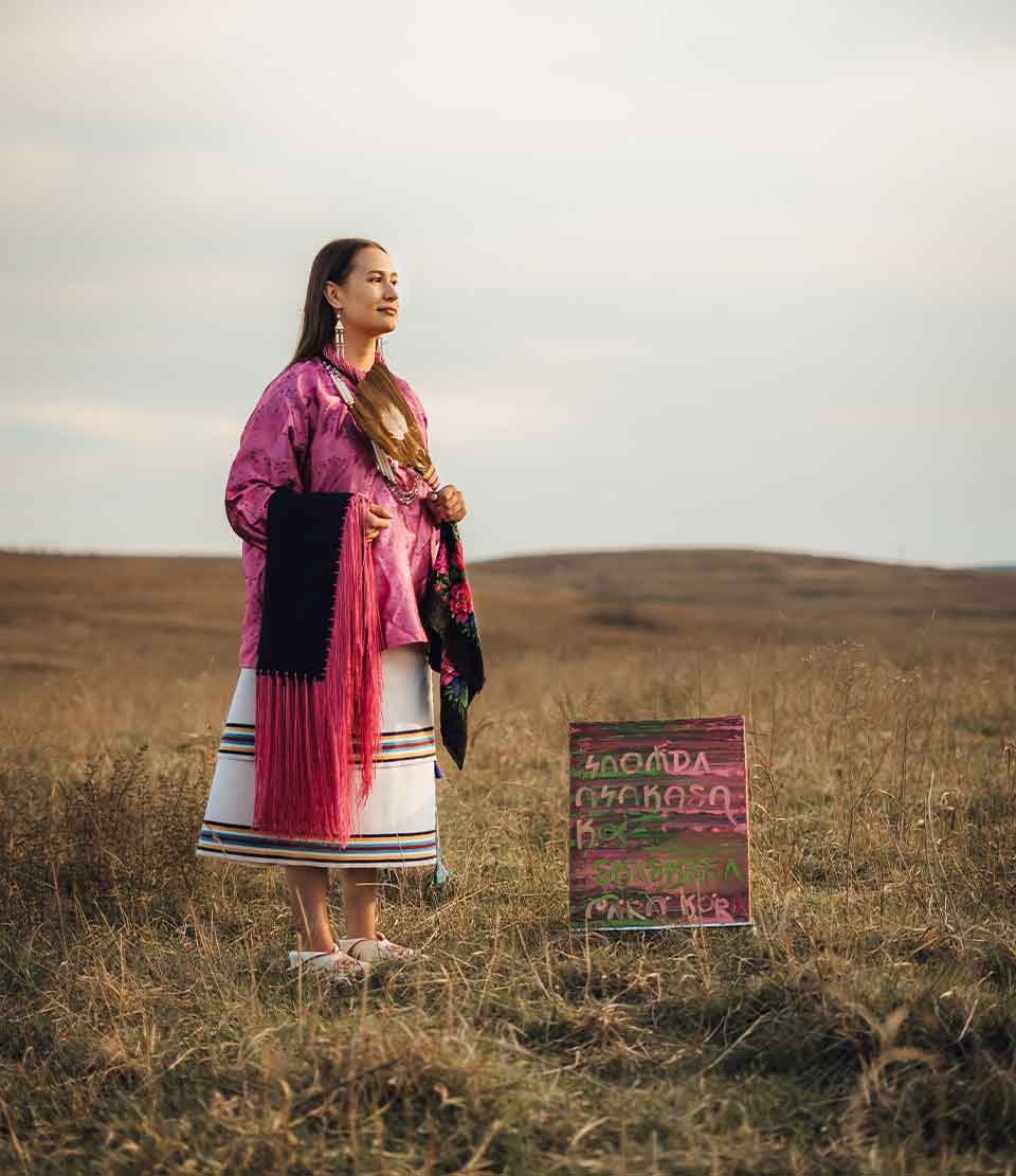 A photograph of a woman dressed in indigenous garb standing on a grassy hilltop. A piece of art with writing on it sits nearby