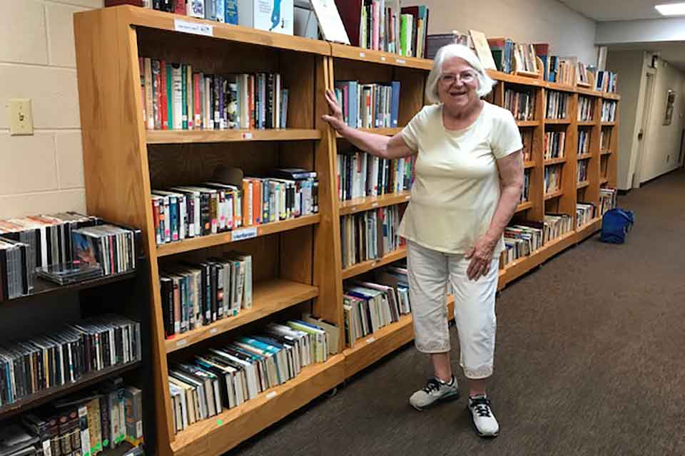 A photograph of a white haired woman standing by a tall vertical bookshelf