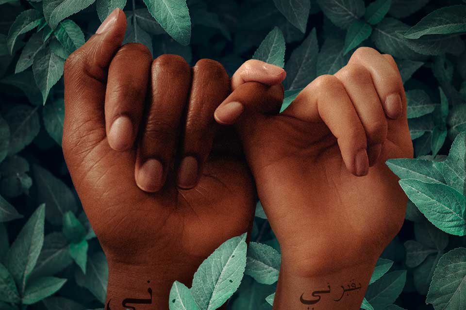 A digital illustration. Two dark skinned hands are bound by a interwoven pinky fingers. Both hands have words in Arabic written on the wrist.