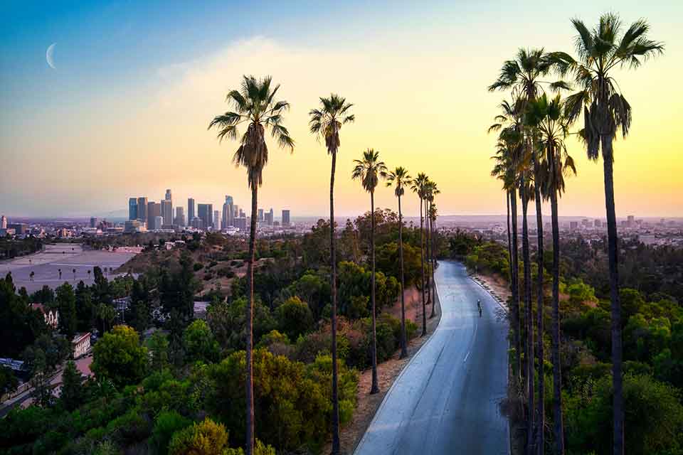 A photograph of LA at dusk. A paved road winds through tree dotted hilly landscape