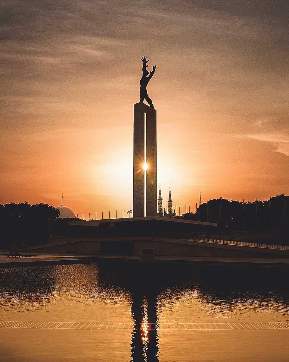 A photograph of a statue of a humanoid figure reaching up on top of a tall, slender tower. The figure is haloed in a rising/setting sun. The tower is reflected in the pool of water in the foreground.