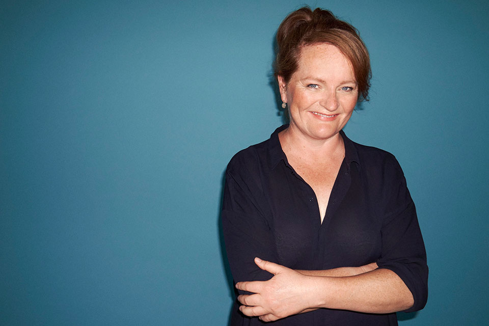 A photograph of Dorthe Nors, who smiles at the viewer, arms folded, against a dark blue background