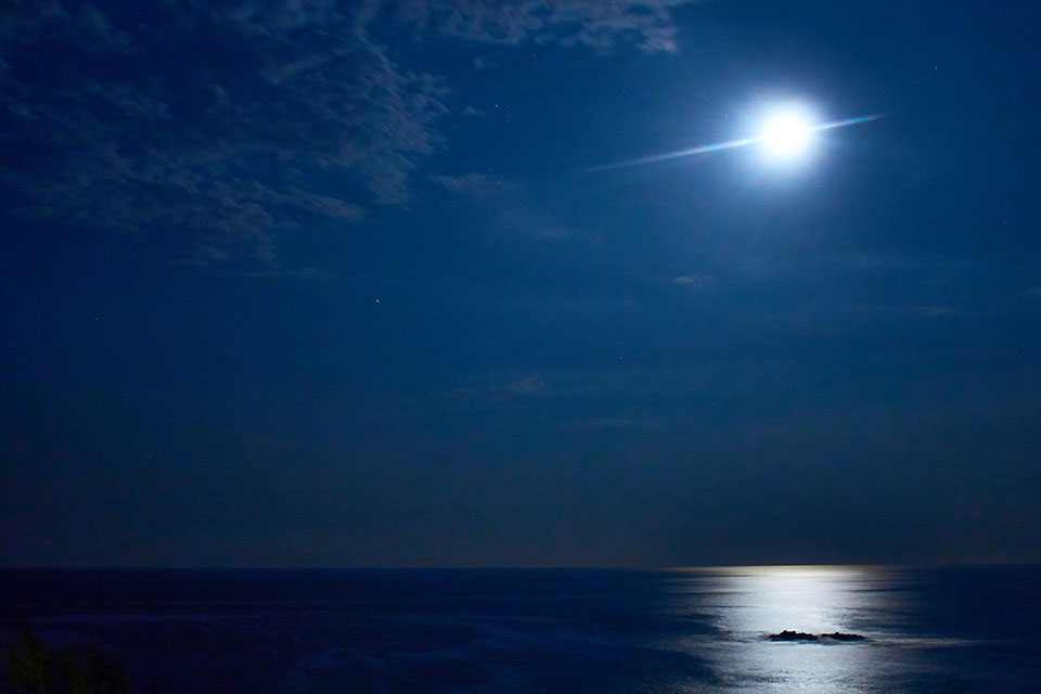 A photograph of a look out at sea, where the moon, just wrapped in gauzy clouds, hovers over a motionless sea below