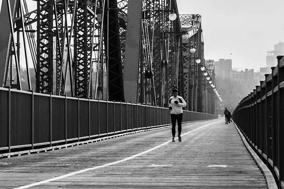 A black and white photograph of a person jogging toward the camera on a long suspension bridge