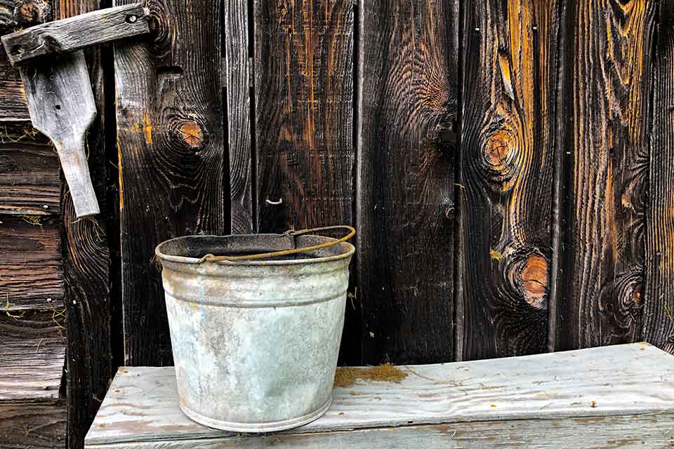 A photograph of an old galvanized steel bucket positioned in front of a wooden wall